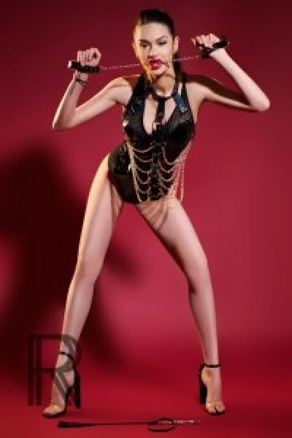 Maddison wearing a decorated latex corset biting onto the chains of cuffs 