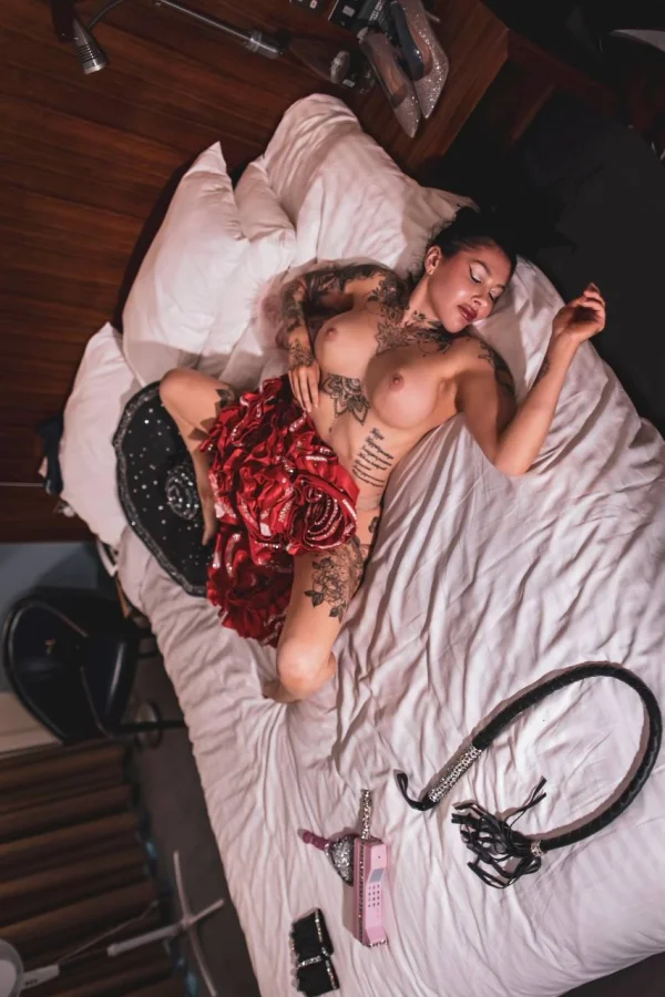 A lady laying on a large bed with white sheets dildos and whips laying on the side 