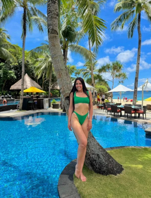 A slim tall brunette escort stood by a large tree with a swimming pool in the back ground 
