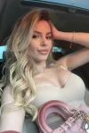 A very beautiful blonde lady has taken a selfie of her in a car wearing a beige dress 